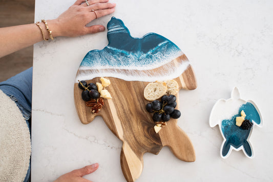 Wooden cutting board shaped like a turtle with Ocean blue resin , placed on a white surface with the matching small ceramic turtle tray 