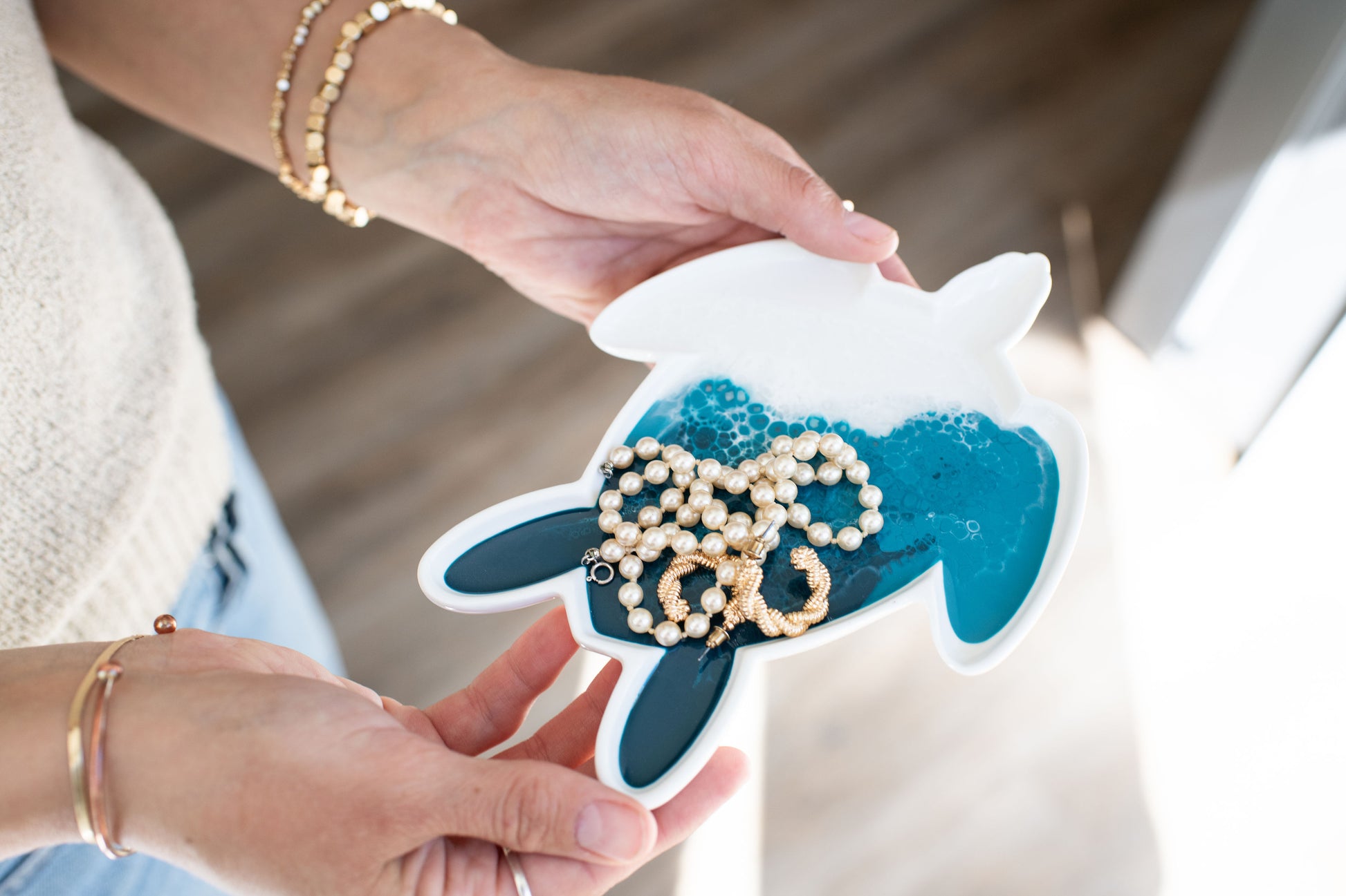 Person holding a white turtle shaped ceramic tray with Ocean blue resin containing pearls and gold rings.