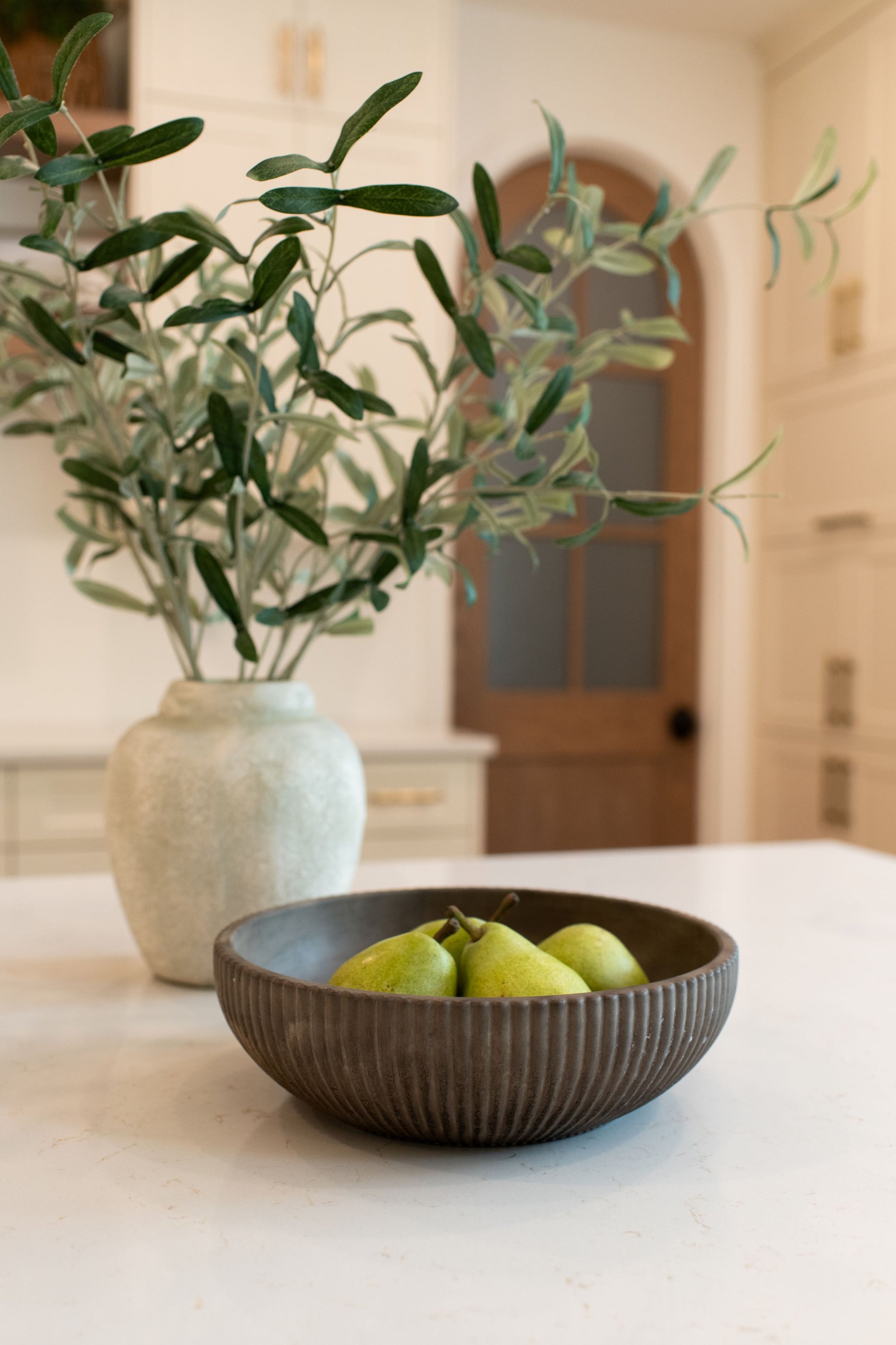 Mocha stone bowl with pears on a kitchen counter next to a vase with greenery