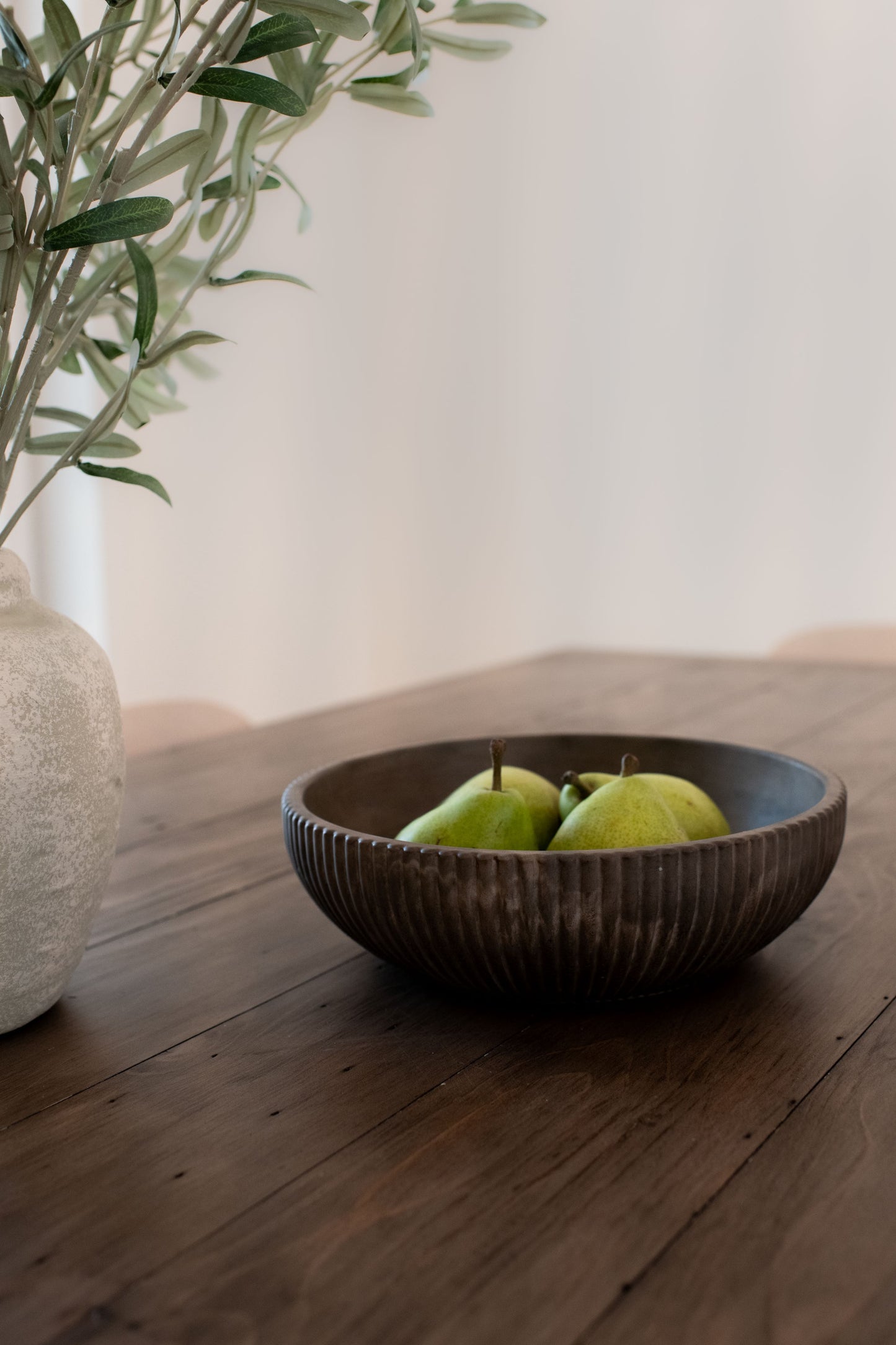 Mocha stone bowl with pears on a wooden surface next to a vase with plants.