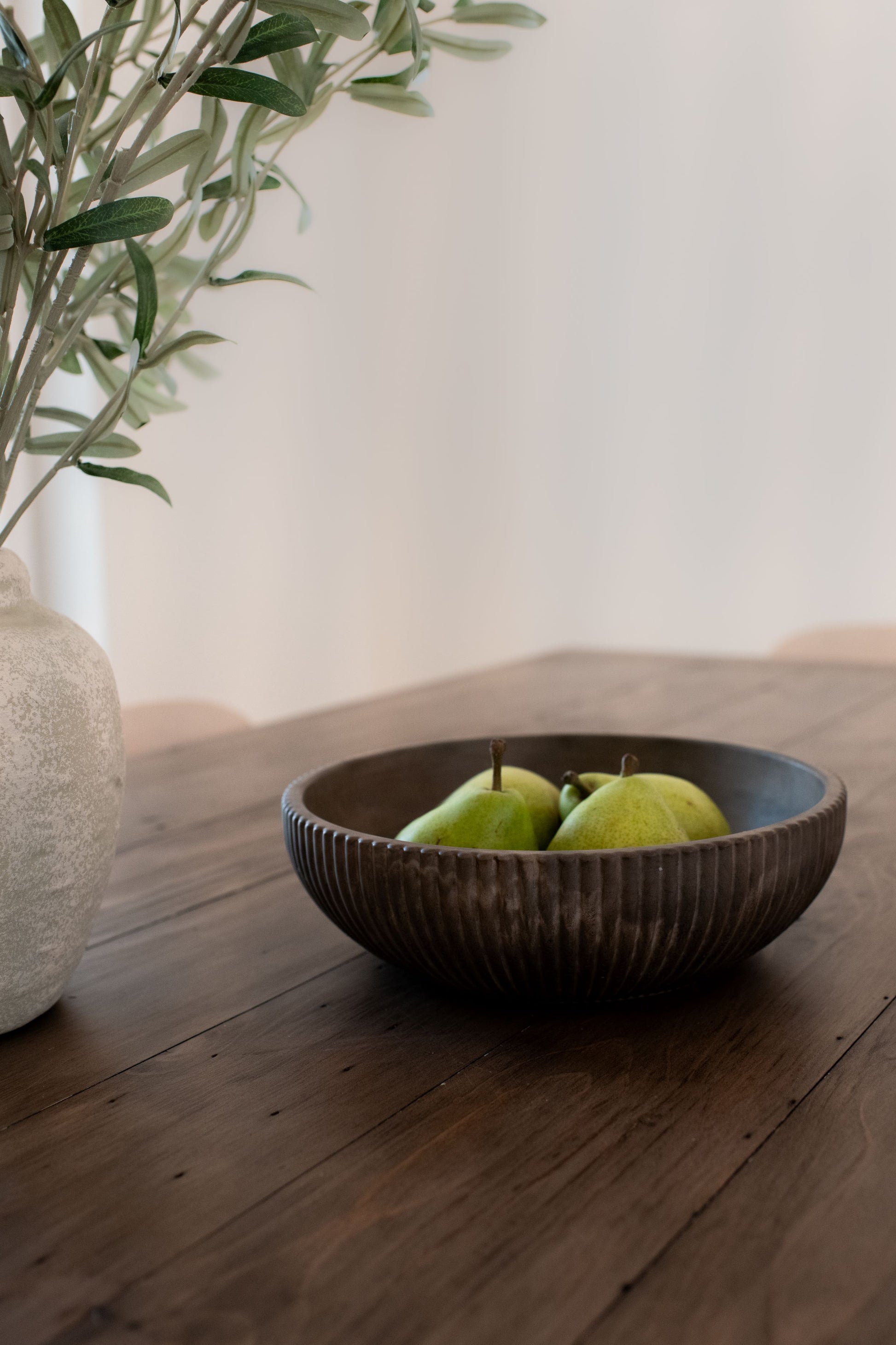 Mocha stone bowl with pears on a wooden surface next to a vase with plants.