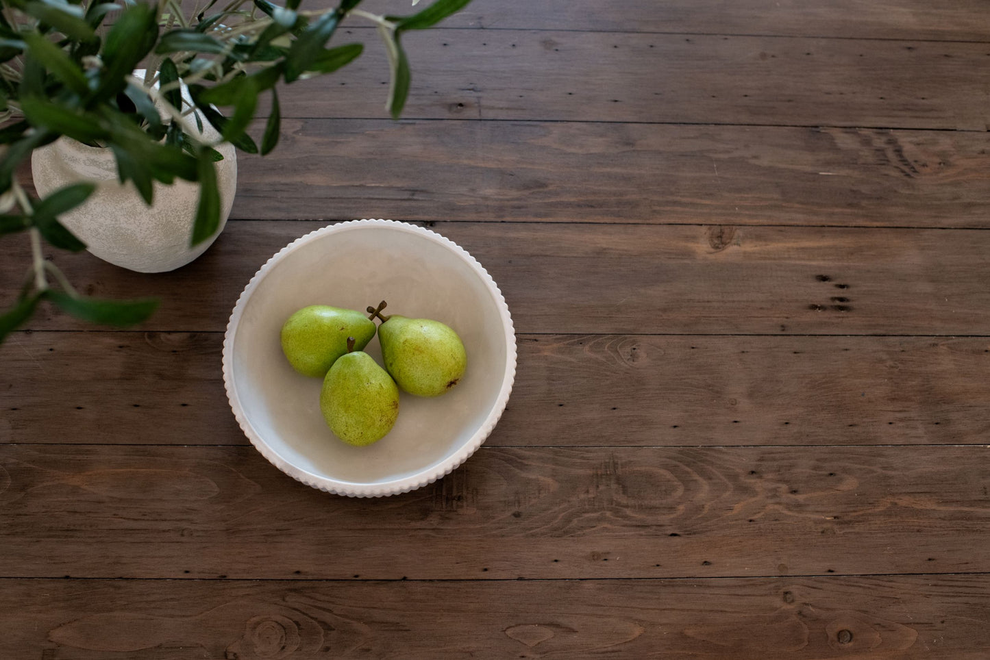 Three green pears in the limestone bowl on a wooden surface with a plant in the corner.