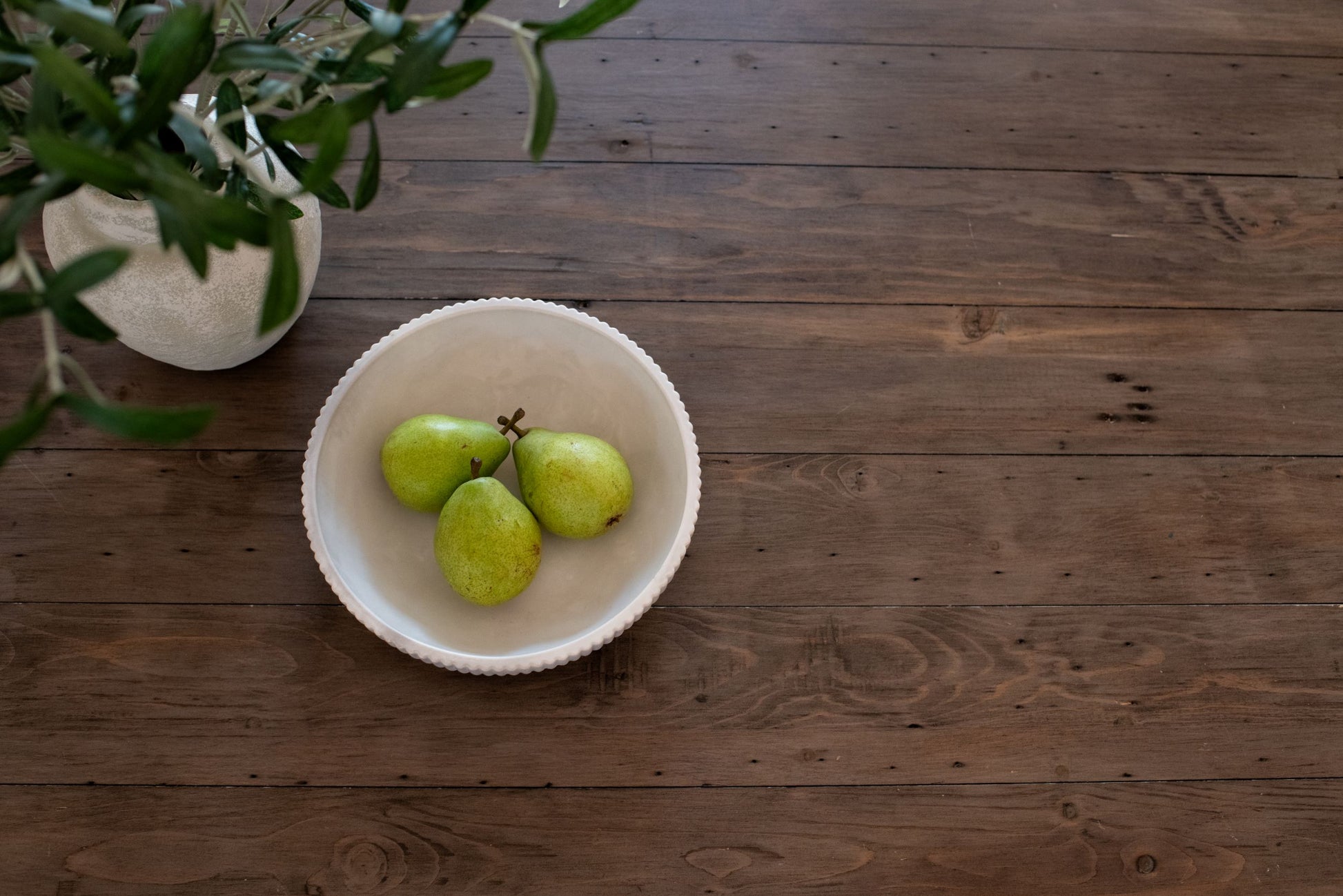 Three green pears in the limestone bowl on a wooden surface with a plant in the corner.